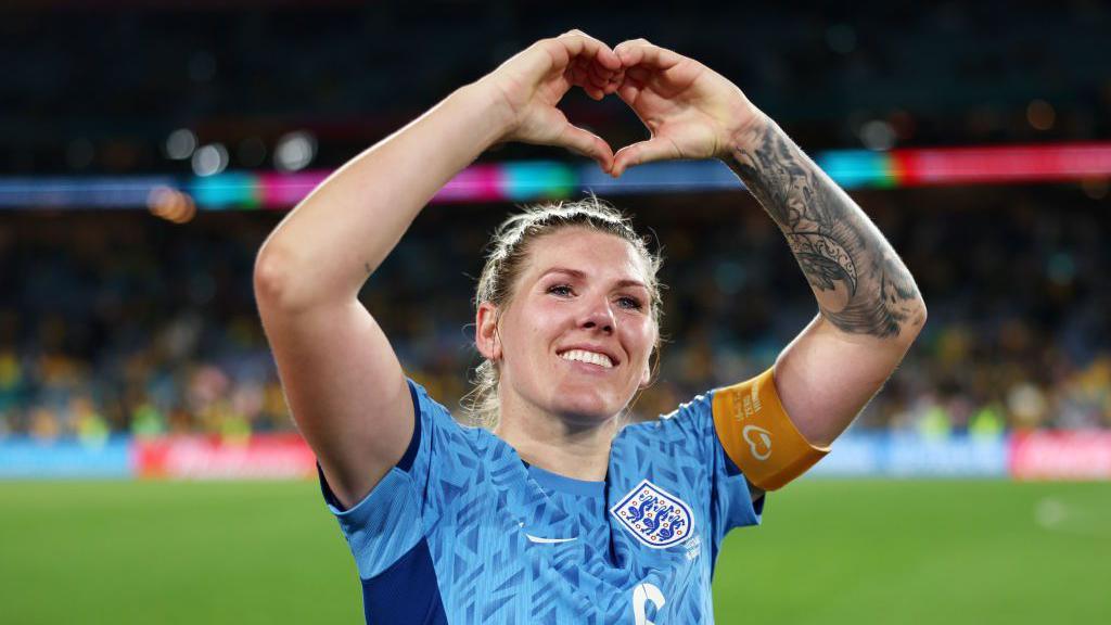 Millie Bright smiling and doing the hand heart sign.
