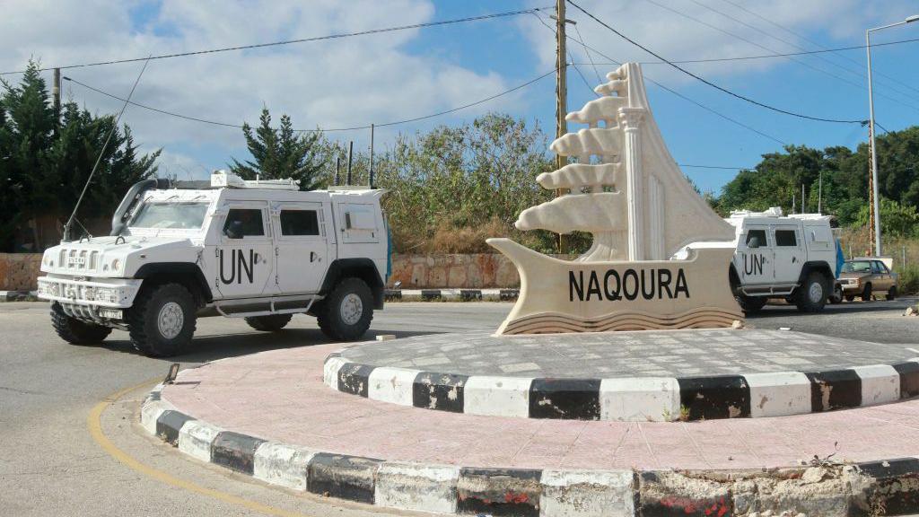 Two UNIFIL armoured vehicles outside the entrance of the southern Lebanese town of Naqoura. They are parked beside a roundabout with a statue with the word Naqoura on it. There are blue skies in the background. 