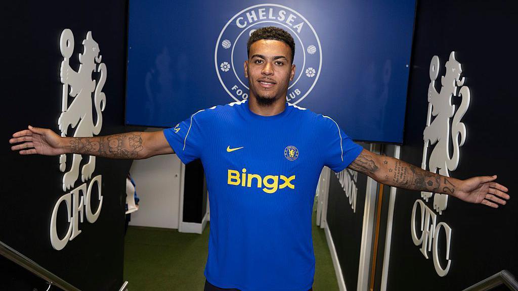Morgan Rogers, wearing a Chelsea kit, stands with arms outstretched inside the entrance to the pitch at Stamford Bridge
