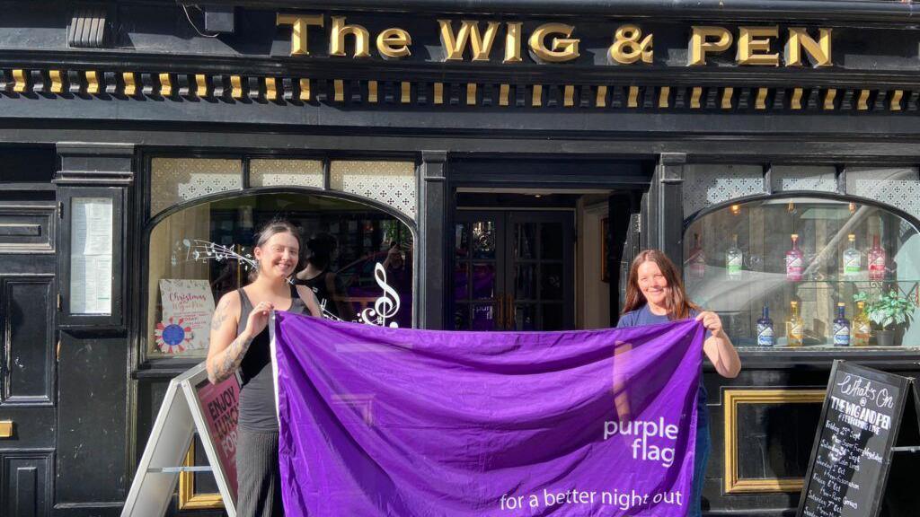 Two women hold up a large purple flag outside a pub.