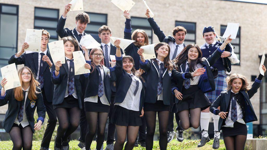 a group of school pupils, boys and girls, all wearing school uniform, jumping in the air while holding a piece of papers.