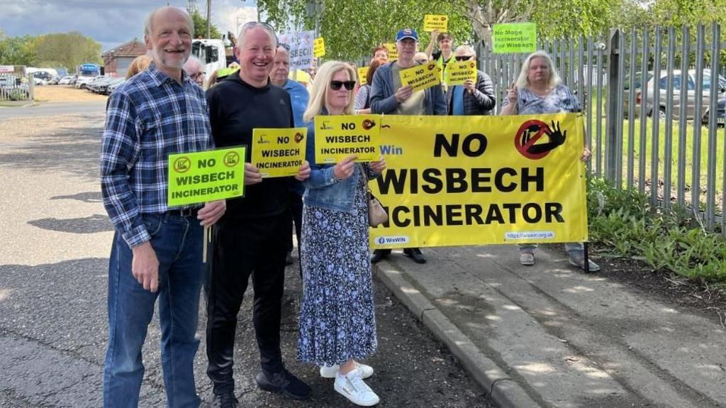 Protesters with signs saying "No Wisbech Incinerator" on Algores Way in Wisbech. They are holding banners which are yellow and green and are standing on a road and a pavement 