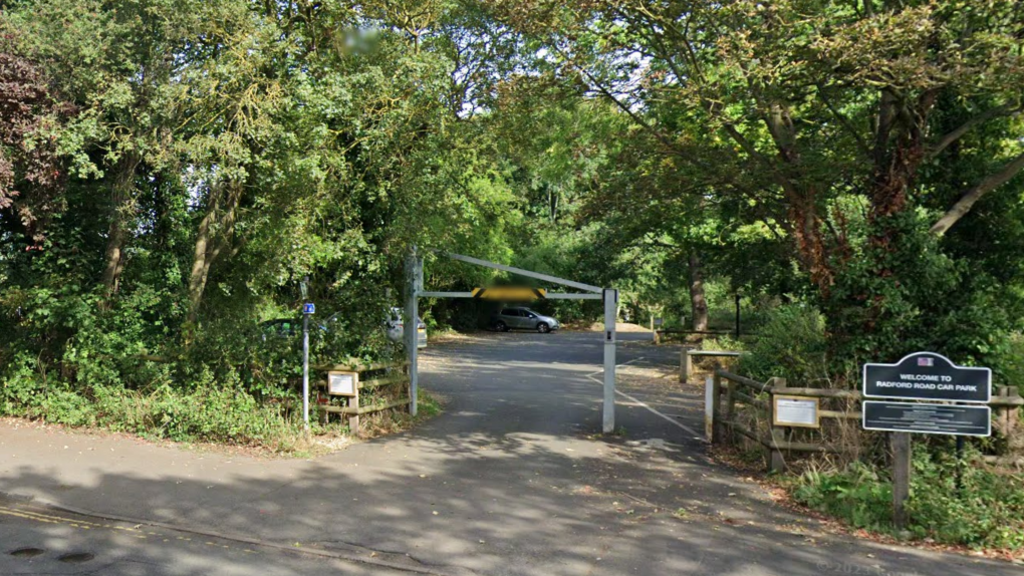 The entrance to a car park, with a height barrier, trees on both sides and a sign saying welcome to Radford Road car park