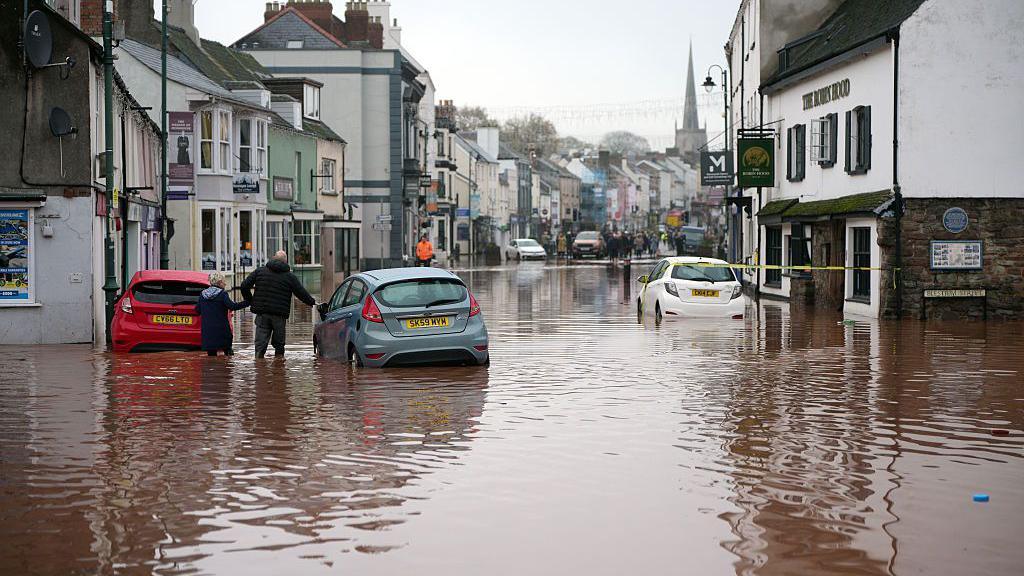flooded street with cars submerged and a few people walking in the floodwater