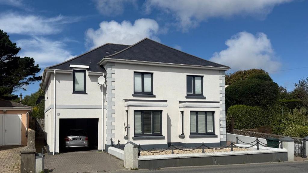 A white house, with a grey car in the garage. There is a road in front of it. The sky is blue with white clouds.
