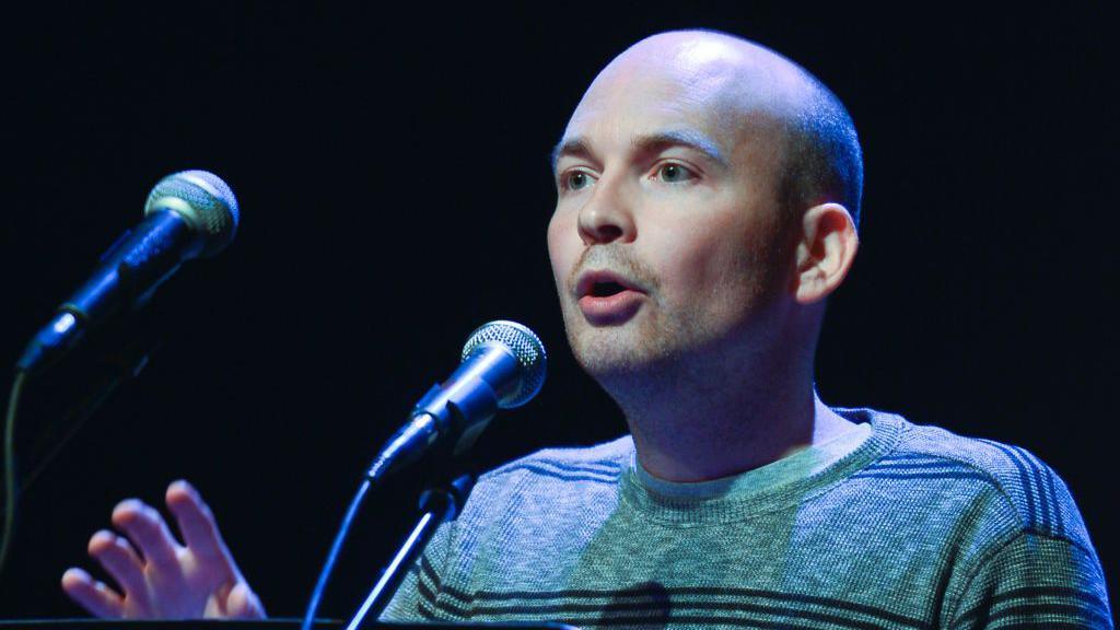 Paul Murphy TD, a bald man, standing in front of two microphones. He is wearing a green top with black stripes on it. The background of the image is black.