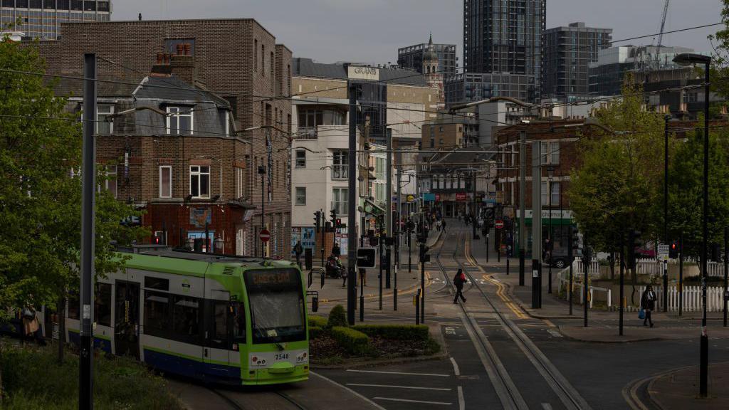 A file image showing a wide view of a green and white TfL tram driving on track through Croydon, south London. Roads, overhead cables, trees, traffic signals, buildings and pedestrians can be seen in the background stretching into the distance