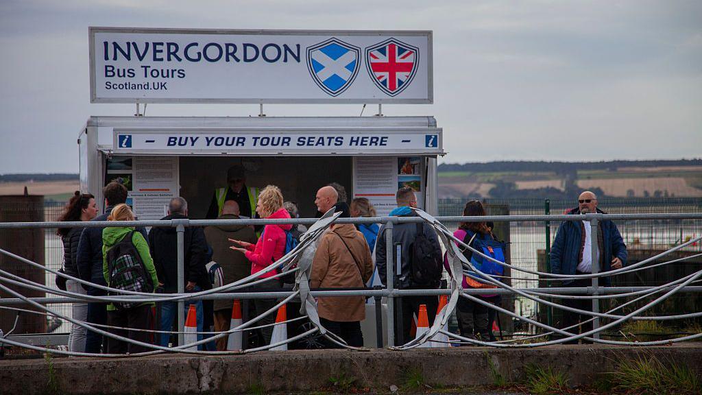 A queue of people outside a kiosk advertising bus tours in Scotland. They are all wearing jackets on quite a grey day. The kiosk is next to the water. 