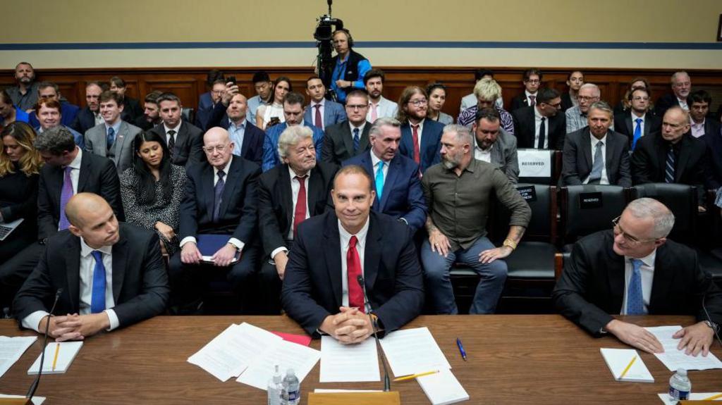 A meeting in a large public room. Three men in business suits are sitting at a large wooden table, papers and pens scattered in front of them. Behind sit ranks of people, variously dressed, with a cameraman at the back of the room.