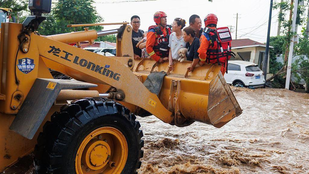 People being lifted above floodwaters by a bulldozer