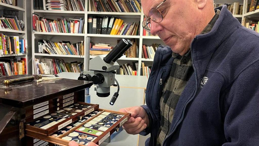 A man with glasses wearing a chequered shirt and blue fleece looks at microscope slides in a small wooden chest