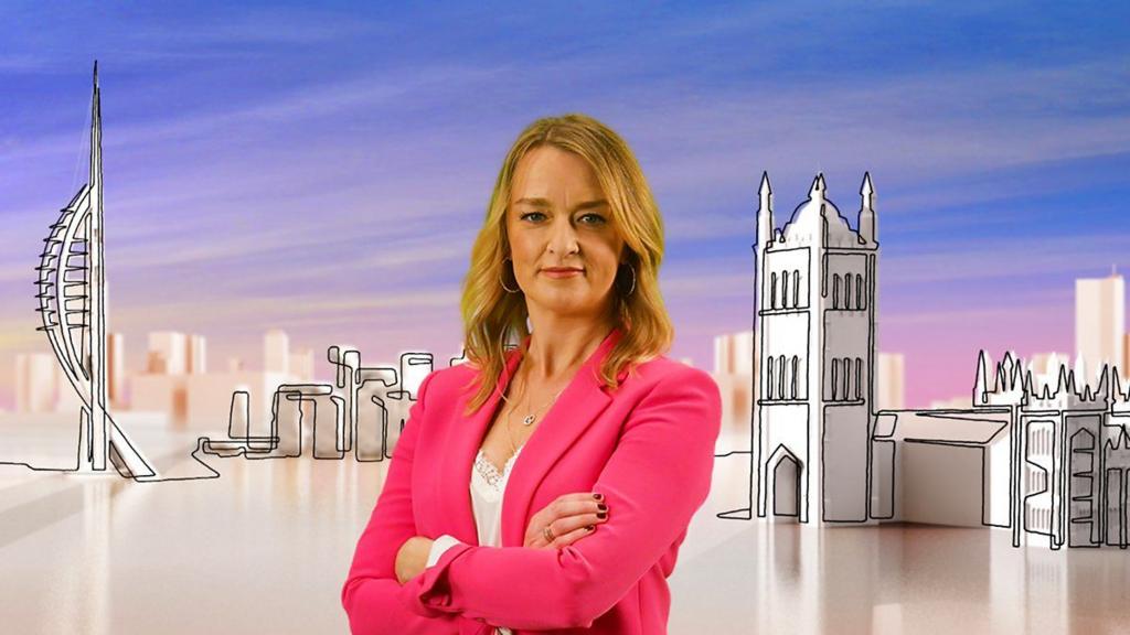 Laura Kuenssberg in hot pink jacket with white blouse with lace at the top. She has her hands crossed in front of her chest and looking directly at the camera, behind her a cardboard model of the House of Parliament to the left