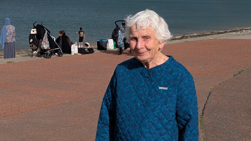 A woman with white hair is seen standing on a beach. She is wearing a blue jumper