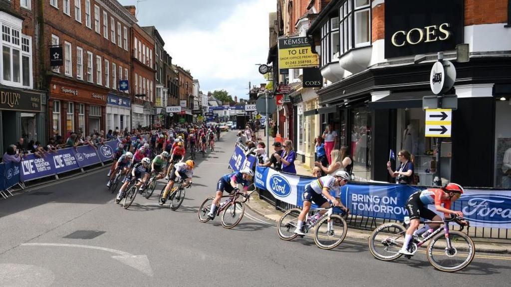 Cyclists ride along Maldon High Street. They are navigating a bend in the road and are in front of shops.