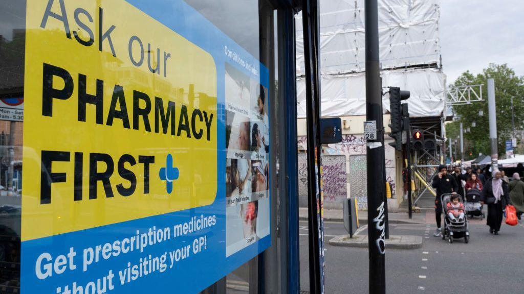 A blue and yellow sign in a pharmacy window advertising the Pharmacy First scheme and encouraging people to ask pharmacists about some common health conditions instead of going to the GP.