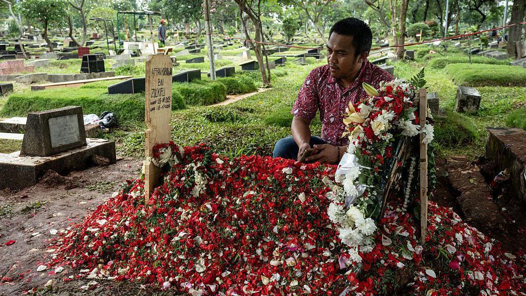 A man holding a mobile phone in his hand and wearing a short-sleeve shirt, looks at the makeshift wooden tombstone of Mr Kurniawan's grave, which is covered in red and white petals