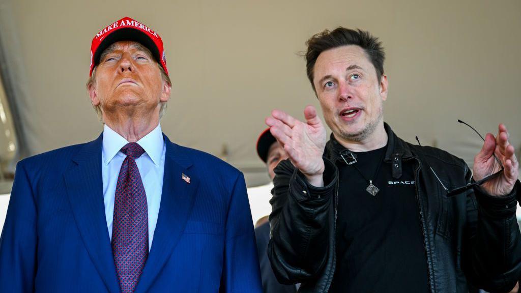 Donald Trump and Elon Musk standing side-by-side. Trump is wearing a blue suit and tie with a red Maga baseball cap. Musk is wearing a black jacket that looks like leather and is holding a pair of sunglasses. 