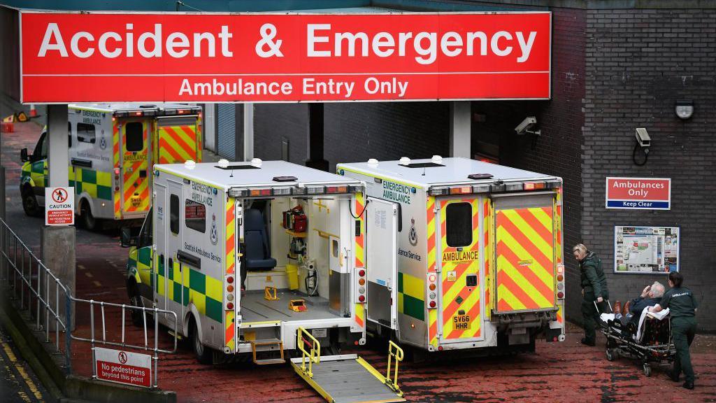 Three ambulances queued up outside the accident and emergency entrance at the Glasgow Royal Infirmary. Two paramedics can be seen dragging a man on a stretcher into the hospital. 