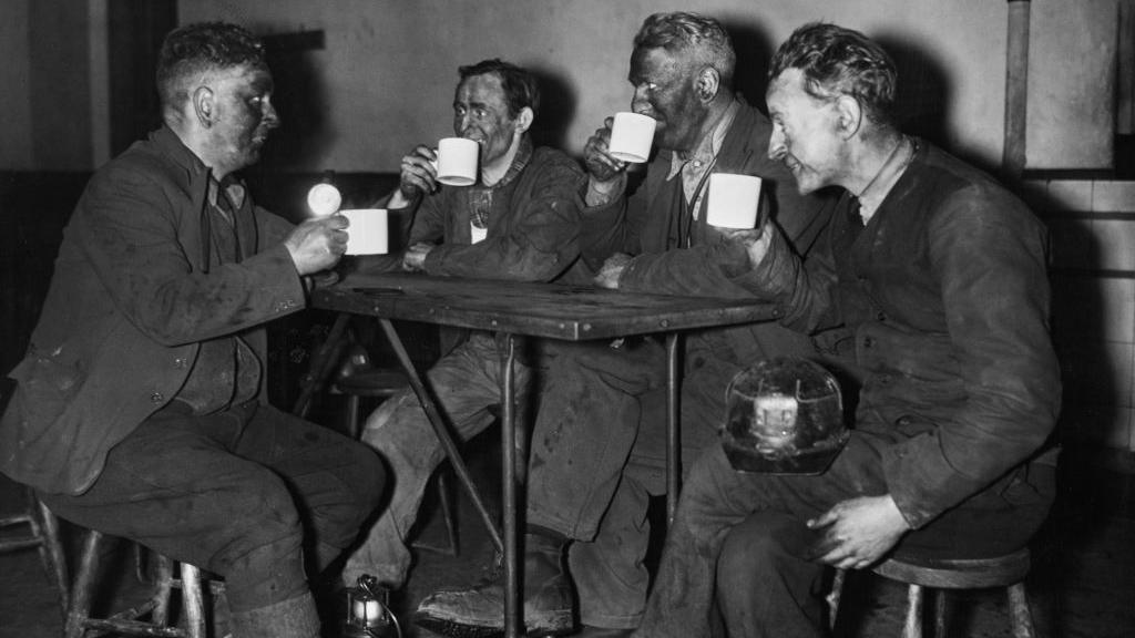 Coal miner Edward Maybank and his workmates enjoy a cup of tea in the canteen of the Merthyr Vale Colliery. It is a black and white photo of four men caked in coal dust wearing blacked overalls. They are sitting around a table supping from pristine white tea cups while smiling.