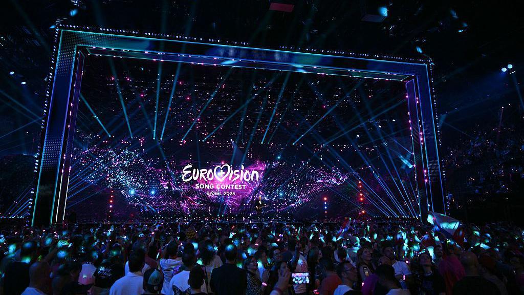A huge stage that is dark, with a screen that says "Eurovision song contest, Basel 2025". There are fluorescent lights of blue, purple and pink. A crowd of thousands stands in front of the stage, reflected with blue light. Many have their phones out.