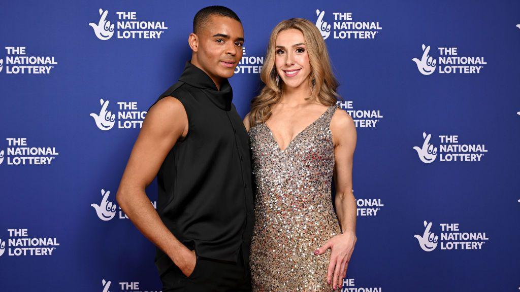 Layton Williams and Lauren Drew standing together at the National Lottery's Big Night of Musicals Red Carpet at the AO Arena. Layton is on the left and is wearing a black suit with one arm in his trousers. Lauren is smiling at the camera wearing a sparkly silver dress with curly light brown hair.