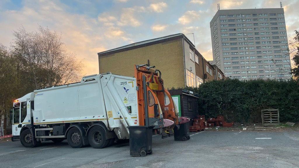 A white bin lorry is parked in an empty concrete car park. Behind it is a hedgerow, a row of houses and a block of flats.