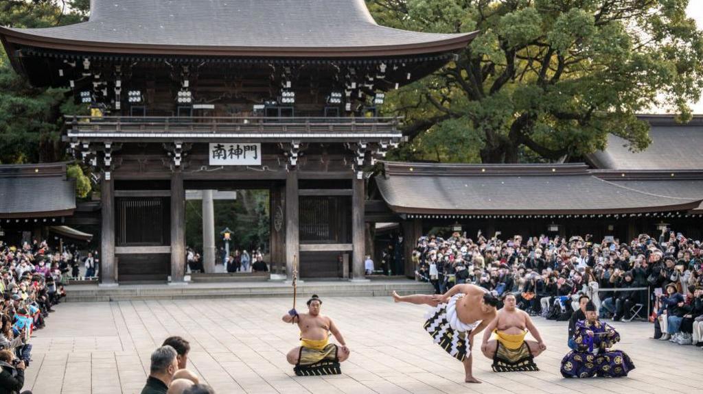 Hoshoryu, topless and in ceremonial dress from the waist down, stamping with a huge Japanese shrine behind. Other wrestlers sit cross-legged nearby and seated audiences are seen in the far left and right of the picture