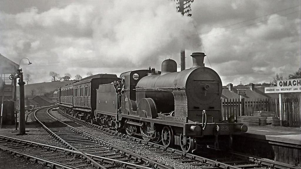 A black and white image of a steam train. The train is arriving at Omagh station as signified by the sign on the right hand side of the railway lines.