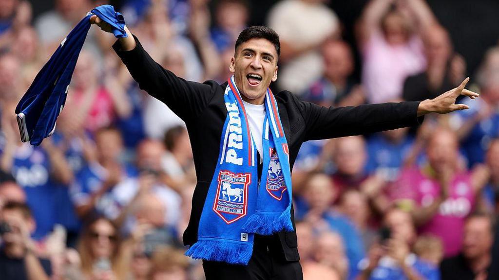 Marcelino Nunez is pictured wearing an Ipswich scarf and holding a shirt in the air. The crowd at Portman Road can be seen applauding him