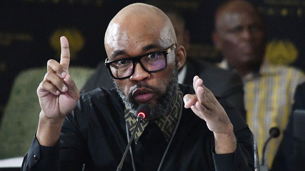 Vusimusi "Cat" Matlala, with the collar of a Fendi shirt sticking out above his jumper, gesticulates as he talks into a microphone.