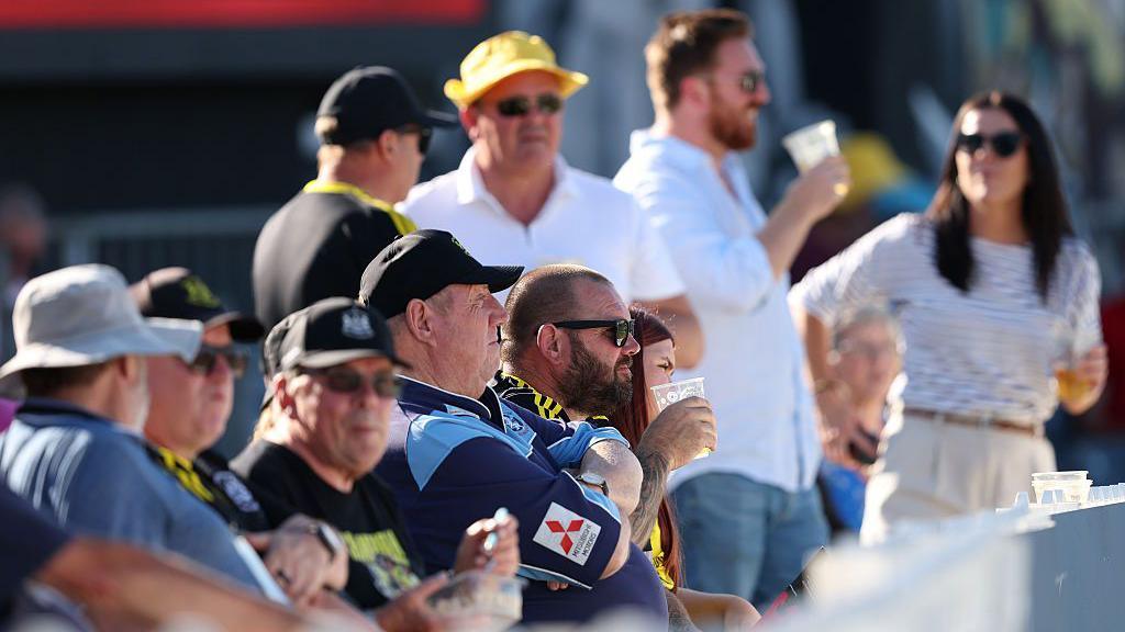 A row of cricket fans seated looking to the right of the camera, many are wearing caps and holding beer in their hands. It is a bright, sunny day.