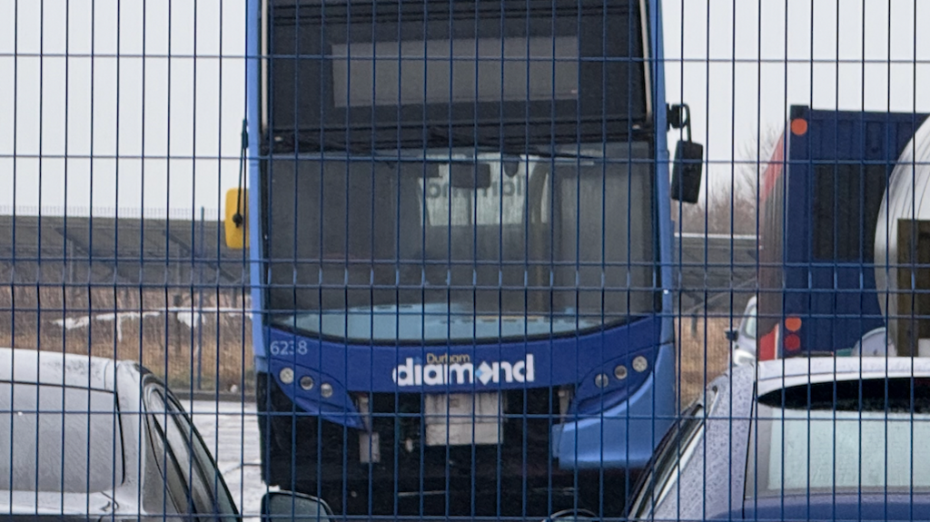 A double decker bus which has been painted in light and dark blue colours, and with white lettering spelling 'Durham Diamond' is parked without some of its front panelling, revealing mechanical parts behind it. It is photographed from behind blue metal fencing.