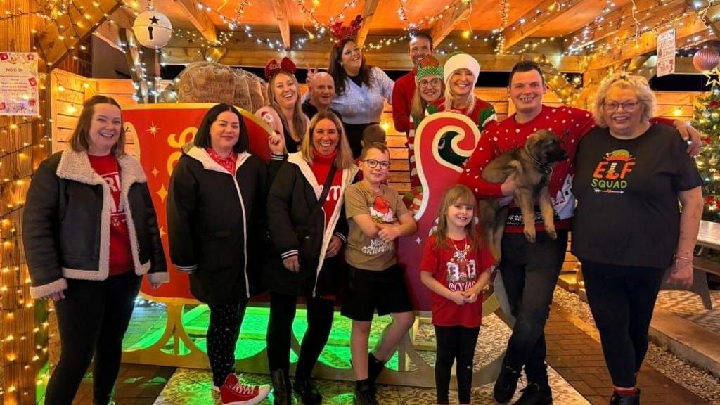 A group of people surrounded by bright festive lights, are standing posing for a photograph with a large, red sleigh.