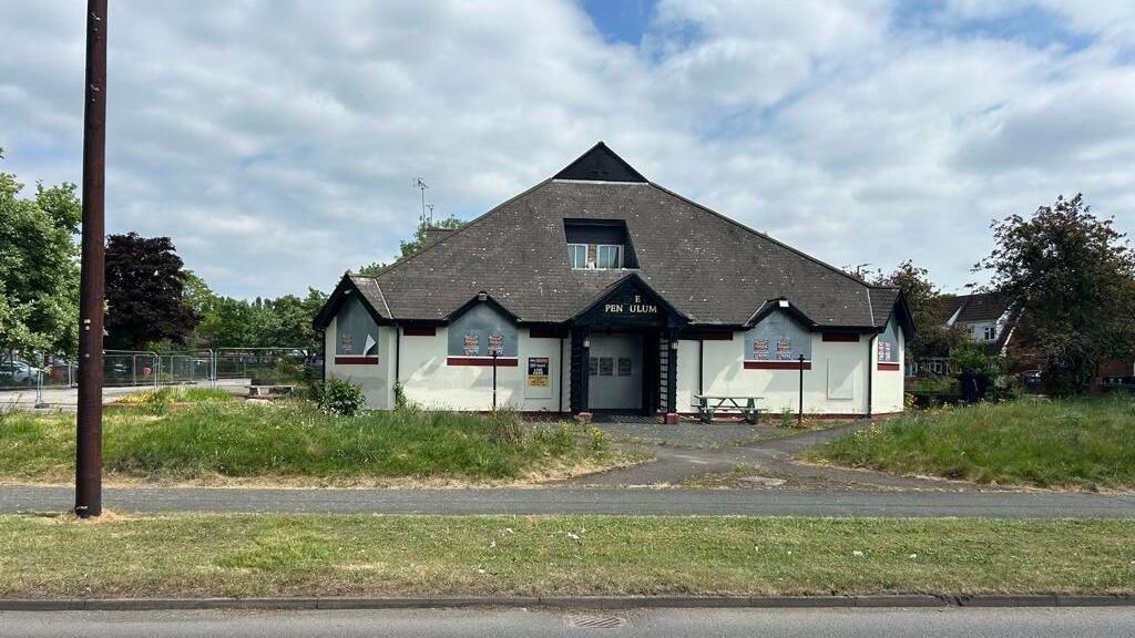 A pub with a large grey triangular roof with grass and a path in front of it.