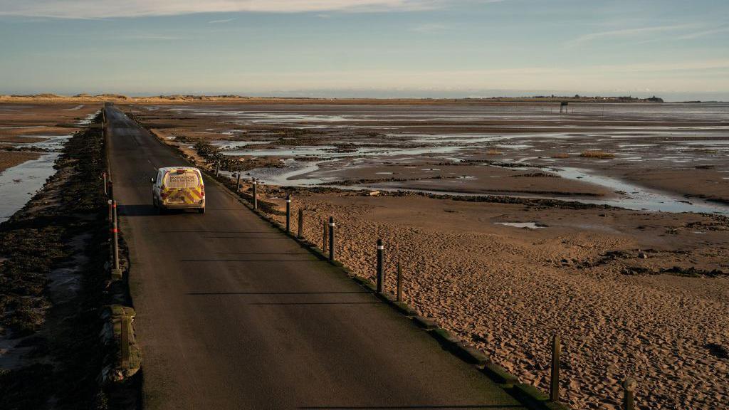 A van travelling across the causeway during low tide.