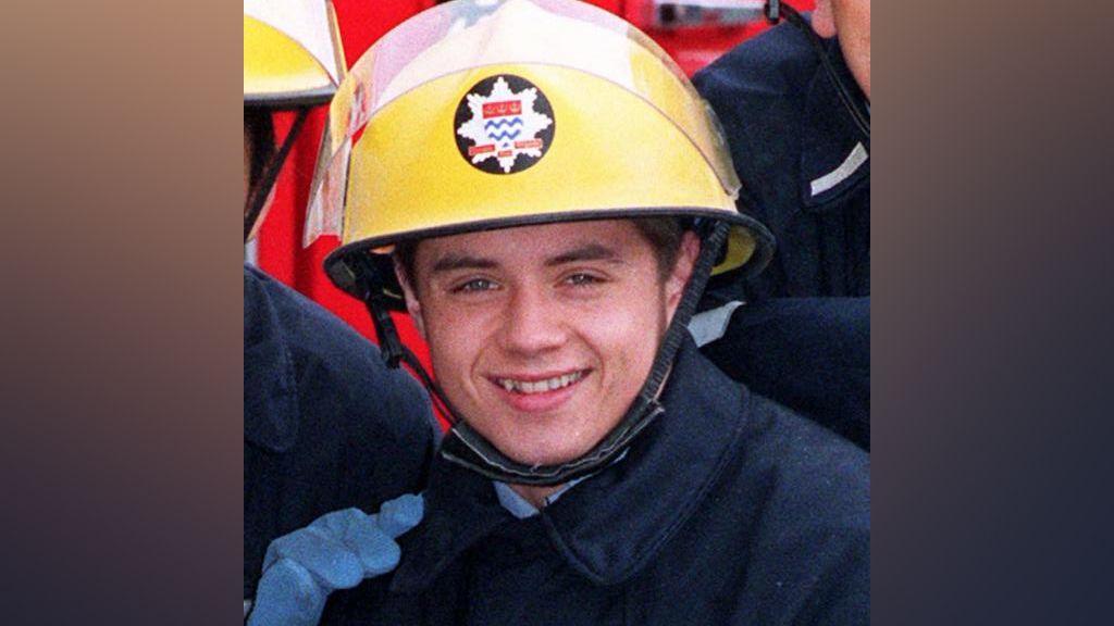 John Alford wearing a fire officer's uniform with a blue jacket and helmet. He is smiling at the camera. 