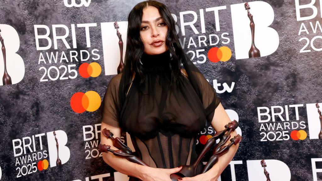 Charli XCX is posing for photographers while holding her five Brit awards in her arms at the awards ceremony.