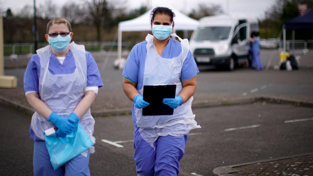 Teo healthcare workers stand in a car park, they are wearing blue medical scrubs and white plastic aprons. They have blue face masks on. One is holding a black clipboard and the other is holding a blue bag.