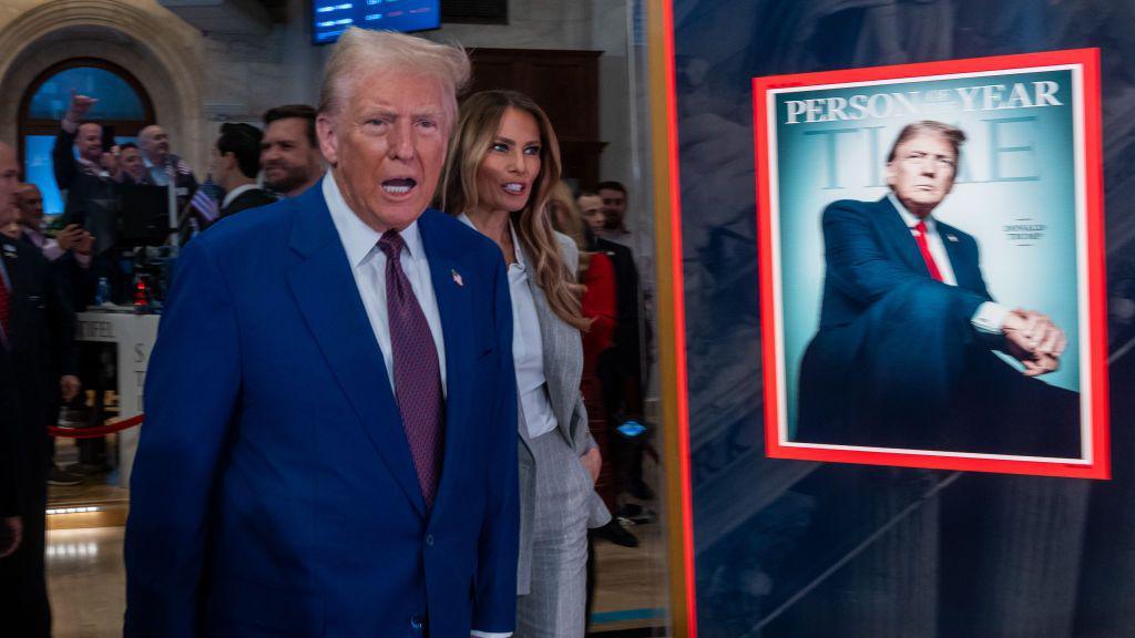 Trump, an elderly man with blonde hair in a blue suit and purple tie, stands next to a Time magazine cover that has an image of him and the words "PERSON OF THE YEAR" on it. Melania Trump, wearing a grey pant suit, stands just behind him