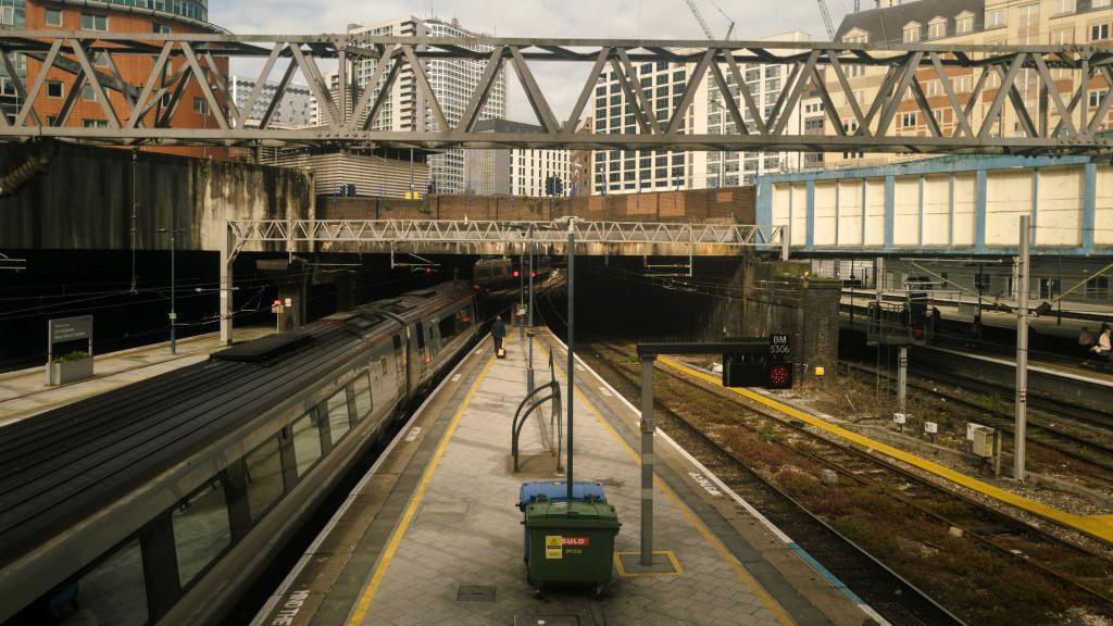 The platforms at Birmingham New Street. There are tall buildings in the background. A train is parked up at a platform. 