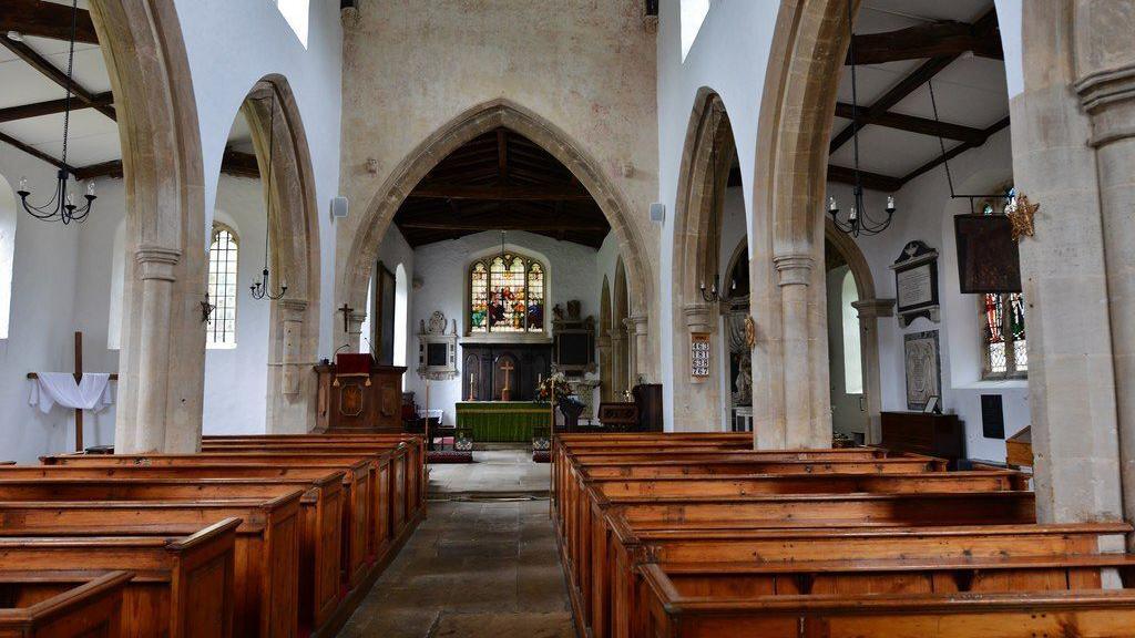 The interior of a medieval church. There are pews on either side and arches. In the distance of an alter with a stained glass window above.