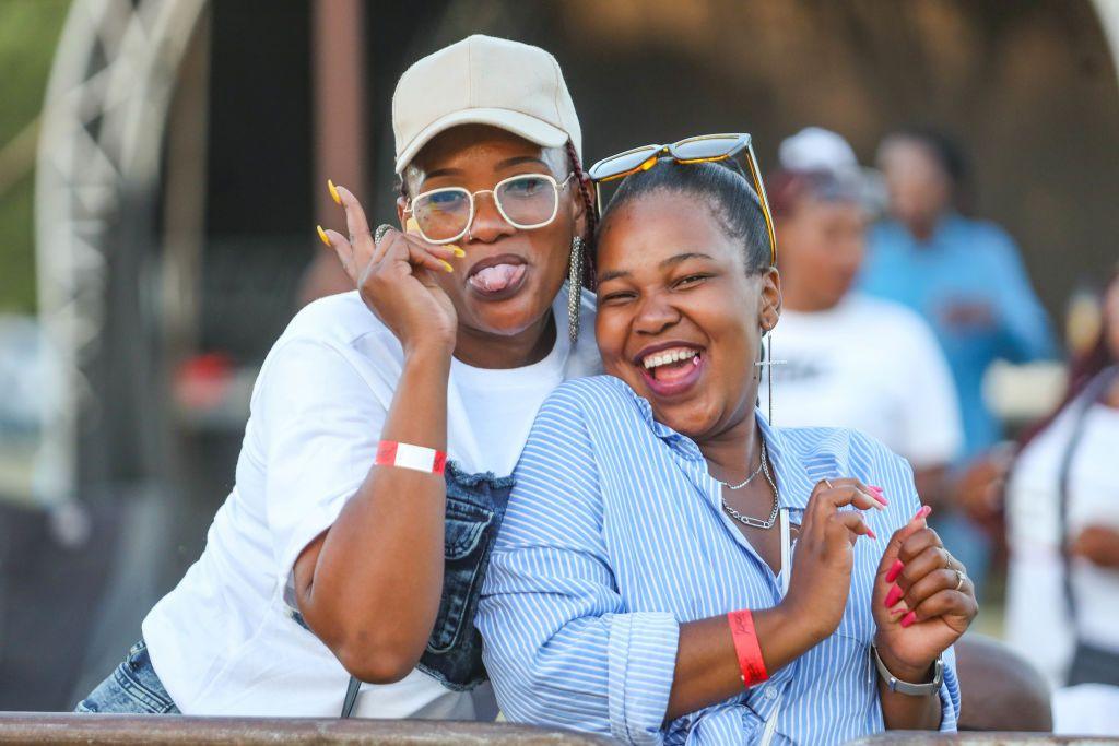 Two women smile and throw peace signs. One sticks her tongue out as she poses for the camera. Behind them is an outdoor stage in Kroonpark Resort - Saturday 7 December 2024