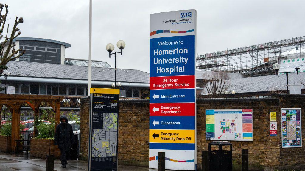 A large sign outside Homerton University Hospital in London displays directions to various hospital services, including the emergency department and maternity drop-off. The hospital building and a construction site with scaffolding are visible in the background. A person wearing a hooded jacket walks past on a rainy day, with wet pavement reflecting the overcast sky.