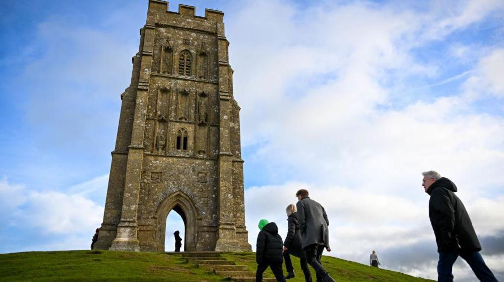 The picture shows people climbing Glastonbury Tor.  The roofless tower of St Michael's Church is in the background.