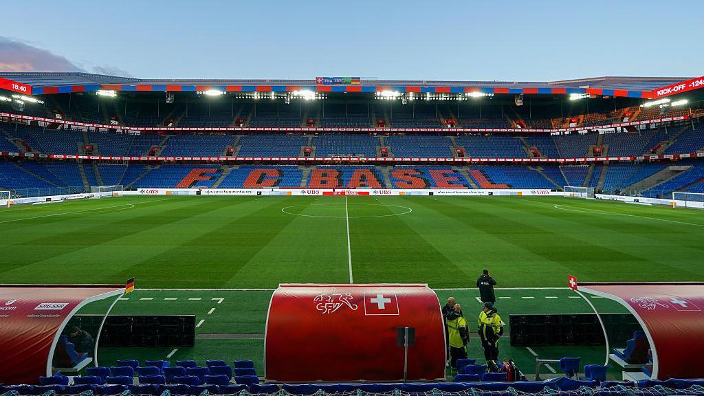 View of St Jakob-Park stadium taken from behind a red dugout on the halfway line, with the name FC Basel in red text on a blue background on the seats opposite