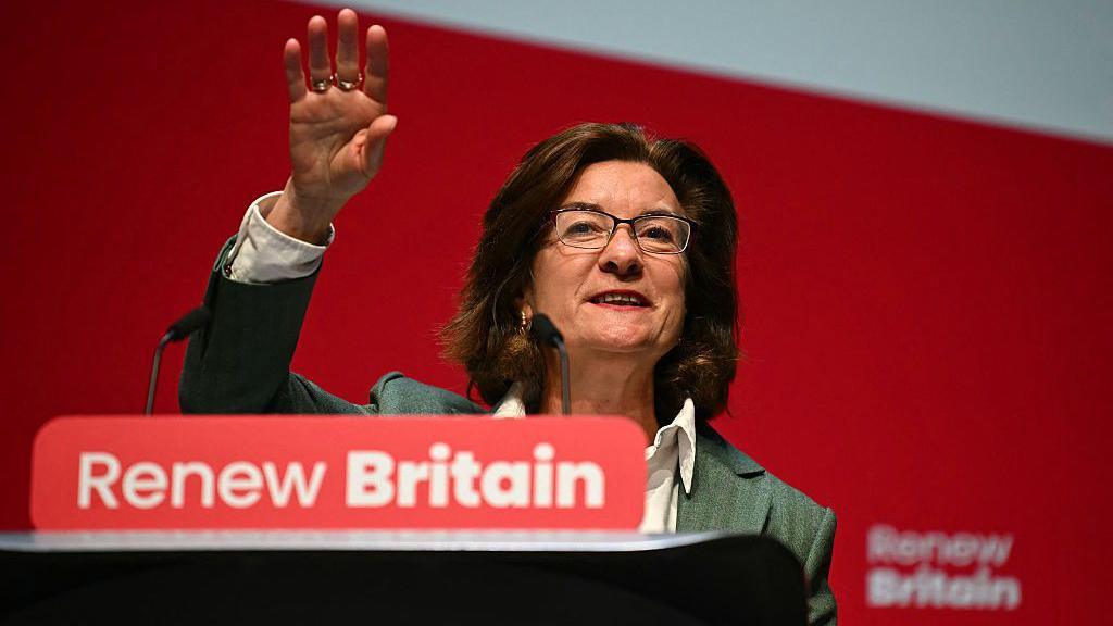 Eluned Morgan, with mid-length brown hair and a white shirt underneath green jacket