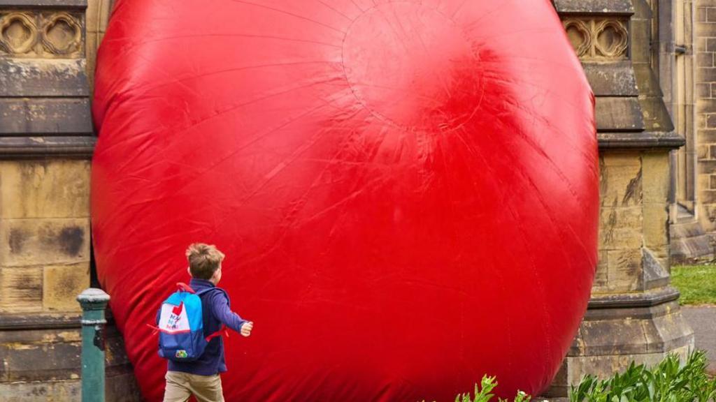A young boy in a blue top with a blue and white bag on his back runs past a large red ball wedged between two stone pillars.