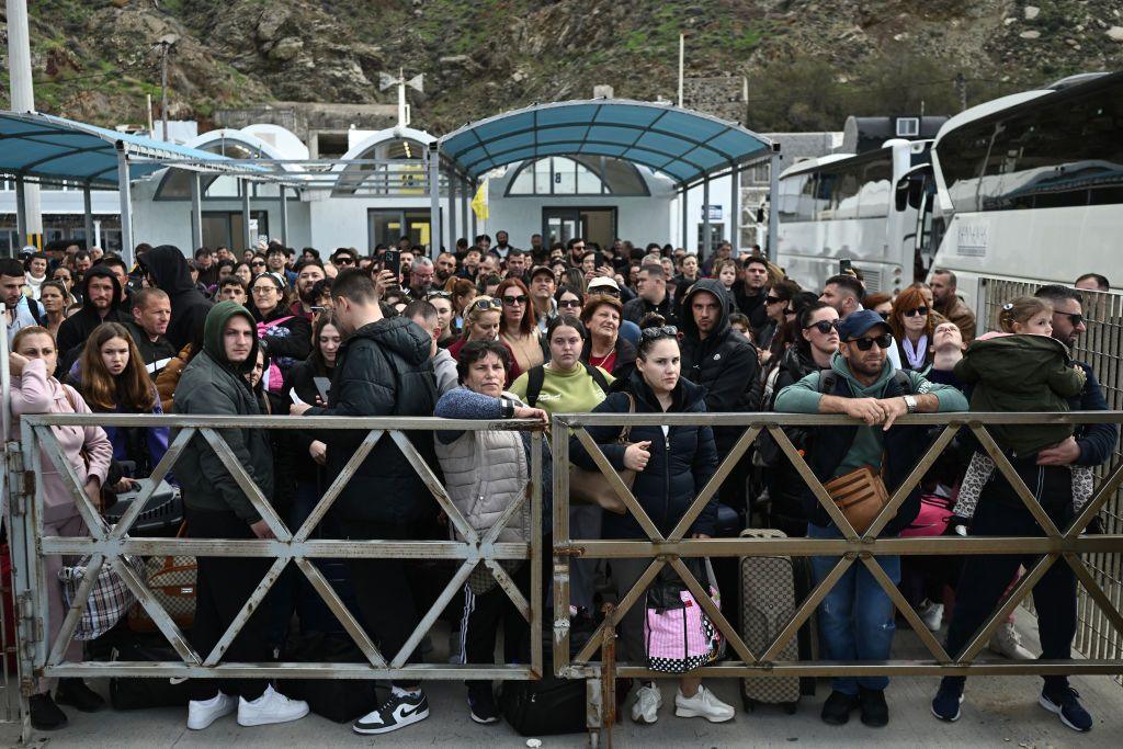 A large crowd of people of a mix of ages carrying bags stand behind a gate. They are stood in front of a ferry terminal and there are large buses next to them