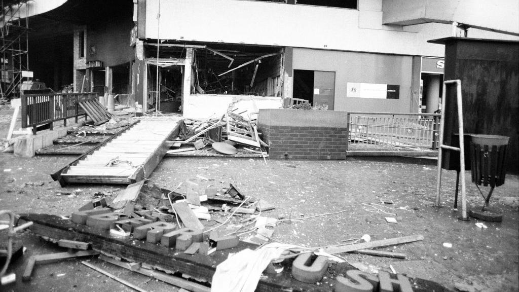 The remains of a pub after a bombing, showing blown out windows on the ground floor, hanging rafters and debris spilling into the street. An external wall sign saying Brew is just visible on a corner leading away from the main door. The pub's main sign lies on the floor in the foreground, broken so only some of the letters can be seen.