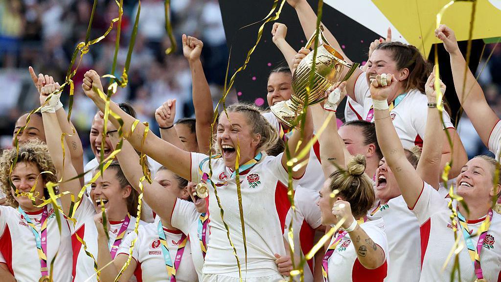 The women of the Red Roses team celebrating their win on the field at Twickenham. They have smiles on their faces, with most raising their arms in the air. One player is holding a gold cup. There are also gold streamers in the air and wrapped around their arms. 